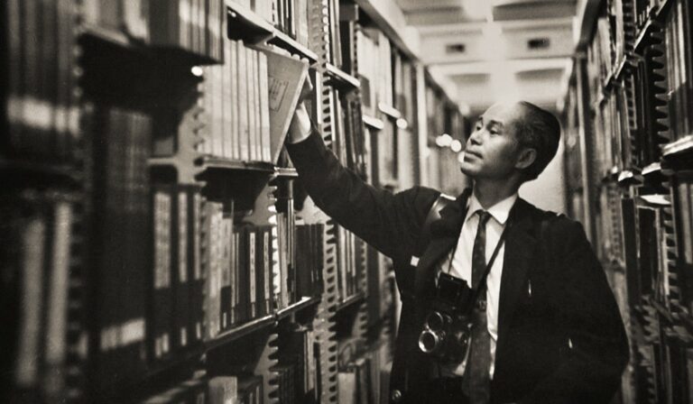 Black and white image of Kem Lee surrounded by tall library shelves, pulling a photo book out of the archives.