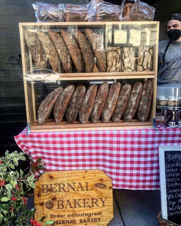 A sign advertises Bernal Bakery and its selection of loaves, displaced in a case on a table with a checkered tablecloth. 