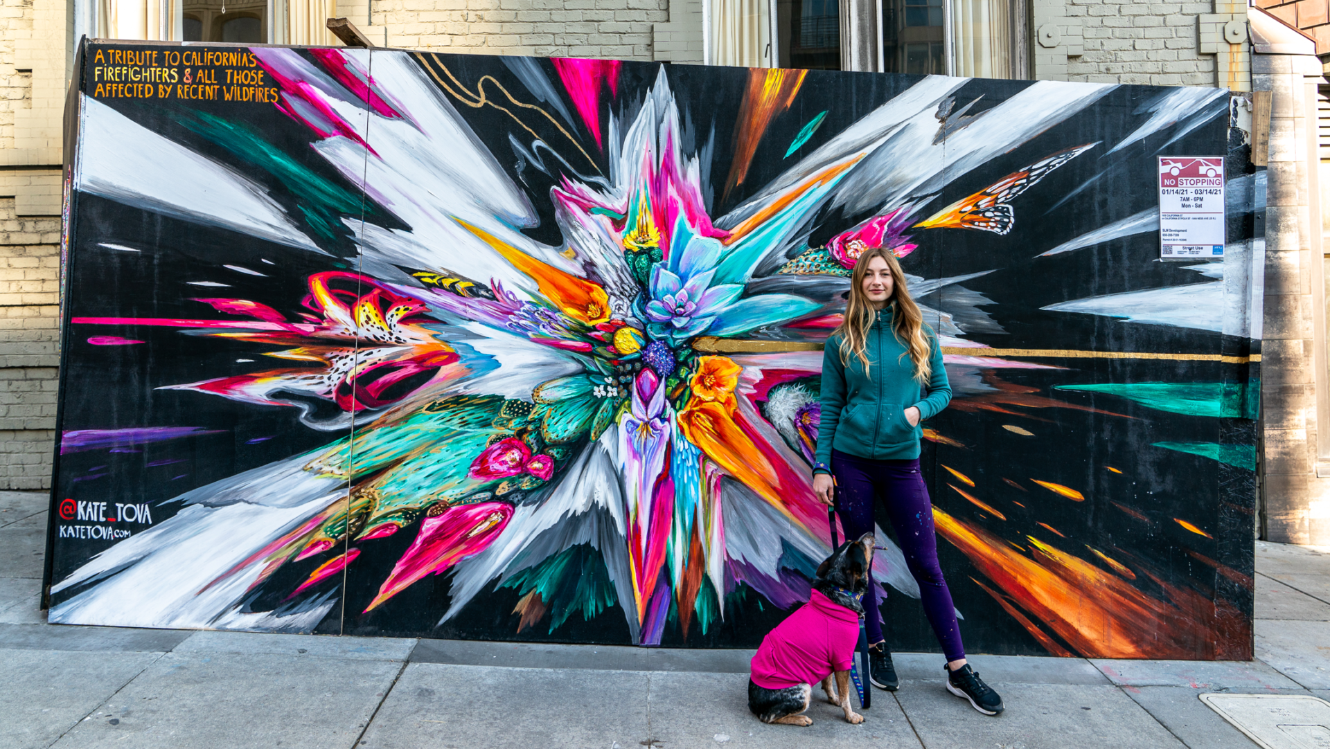 Kate Tova poses in front of her artwok with her rescue dog, Toochi