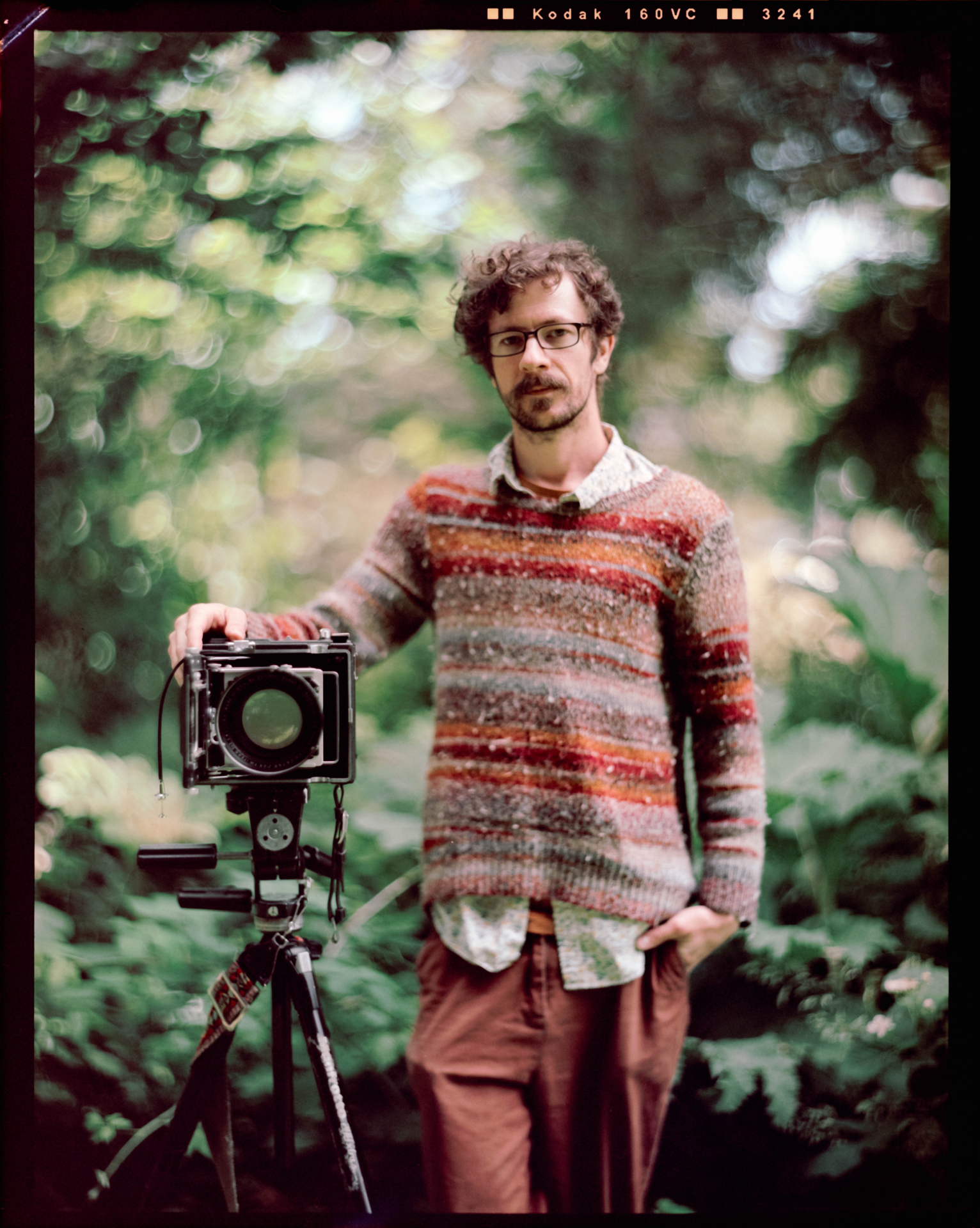 Photograph of Dave, who showed JP the large-format camera ropes, posing next to his camera. Because the back of his camera is open, you can see the green foliage in the background through the lens.