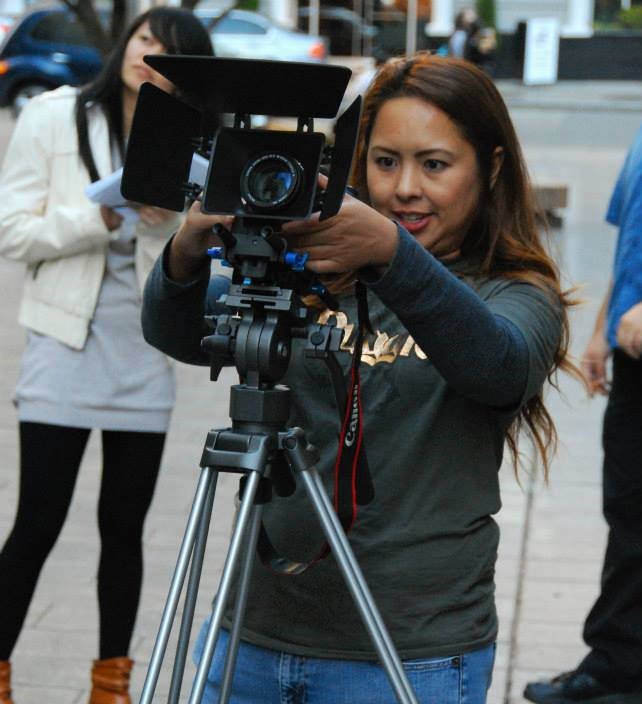 A filmmaker stands outdoors adjusting her camera in preparation for a scene.