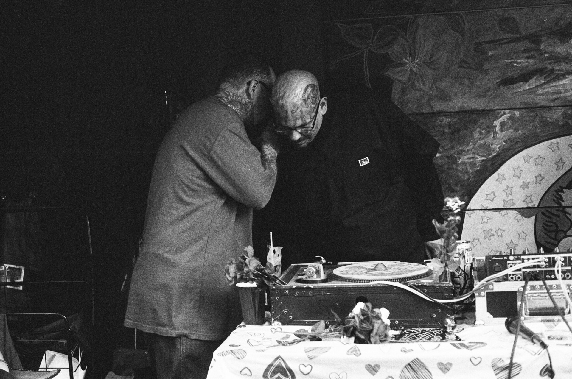 A man whispers something into the DJ's ear behind his record player set-up in Balmy Alley