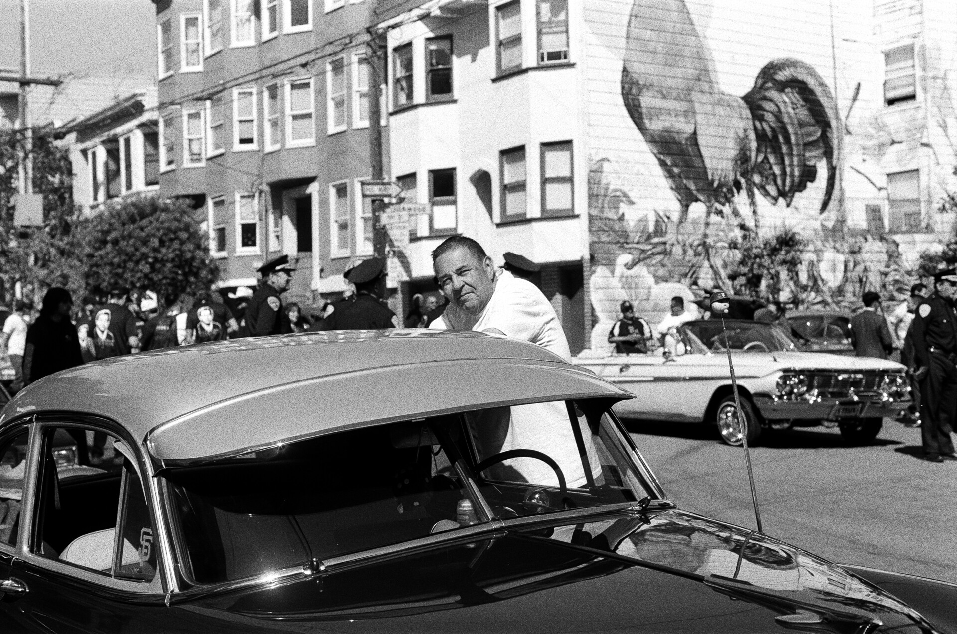 A man leans on the top of his lowrider car, squinting because of the sun