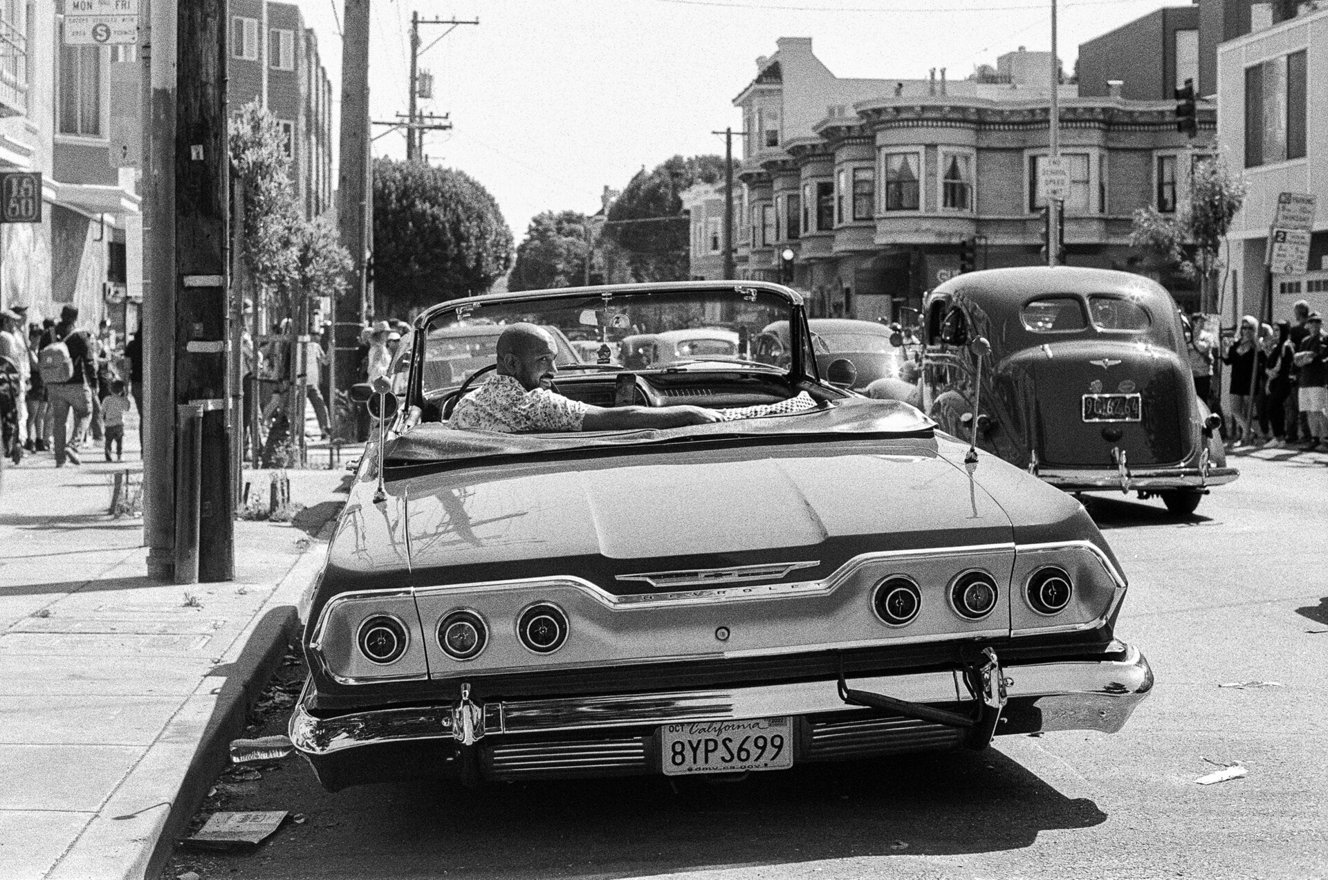 A man looks back over his shoulder from the driver's seat within his convertible lowrider car, smiling for the camera 