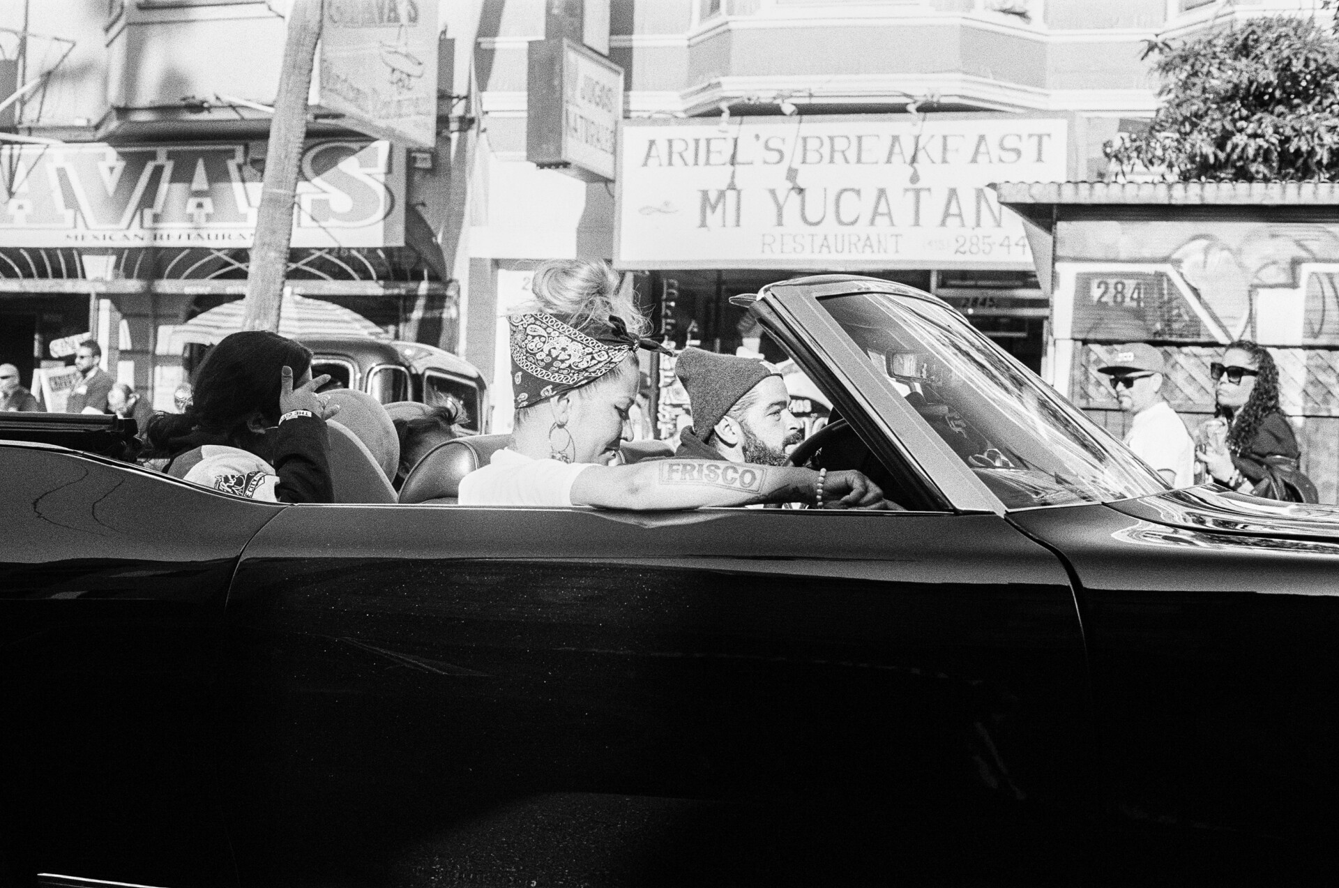 Three passengers ride in a low-rider, including the woman in the passenger seat with arm resting on top of the convertible with a visible Frisco tattoo