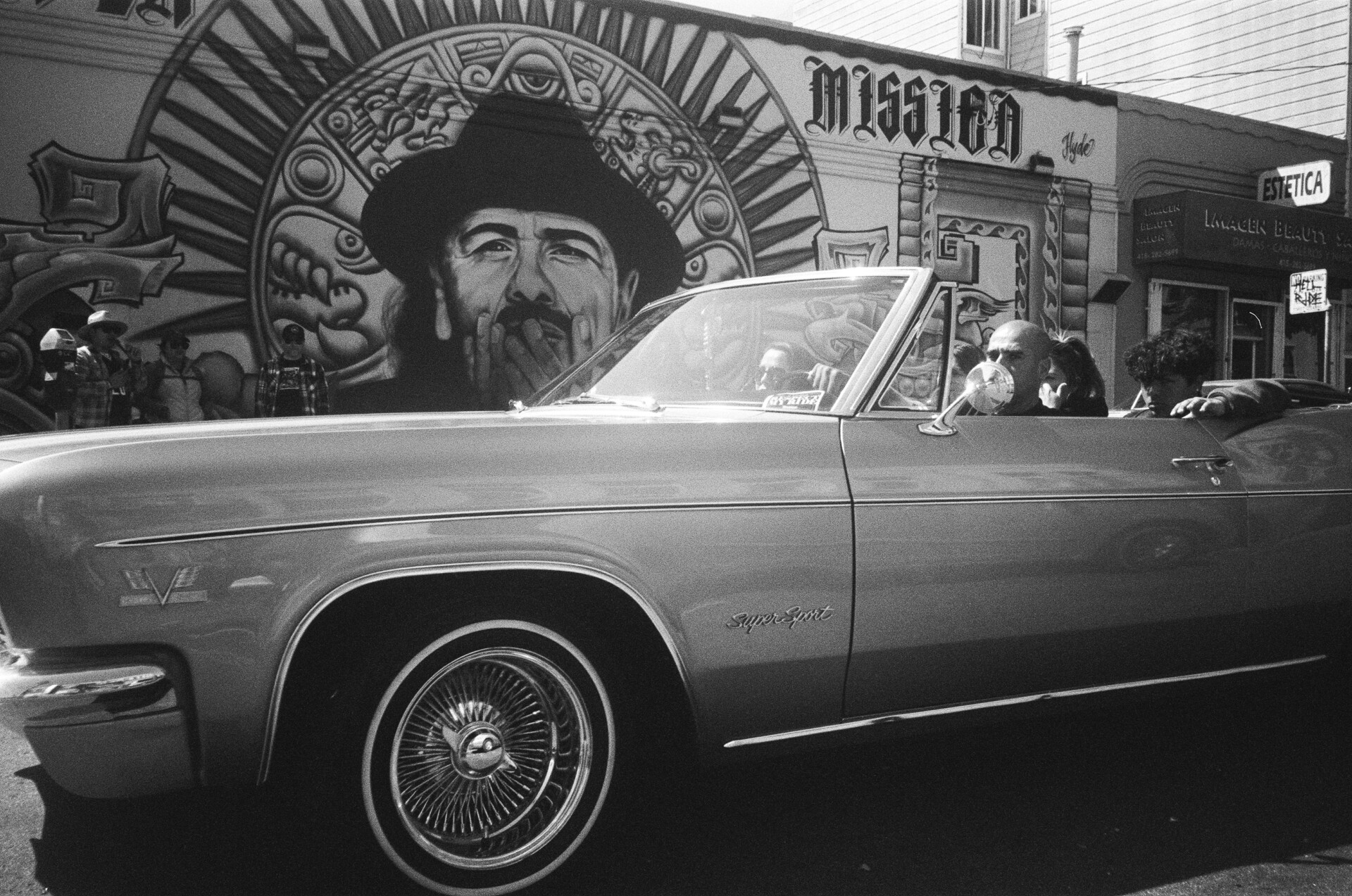 A packed lowrider car drives past a painted wall with a man's face and the word "Mission" painted on it