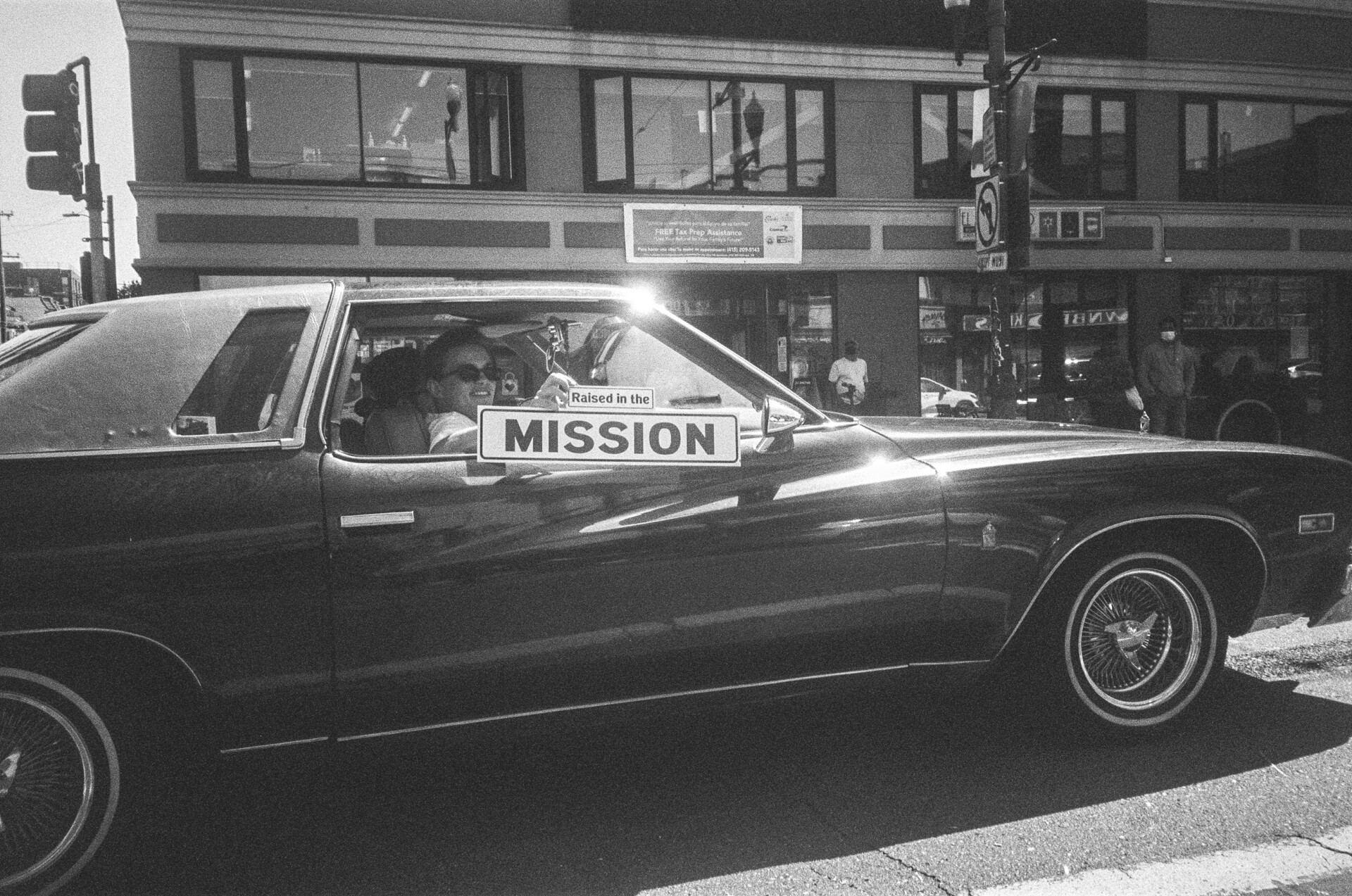 A woman proudly holds a Mission street sign outside of the passenger window of a lowrider car.